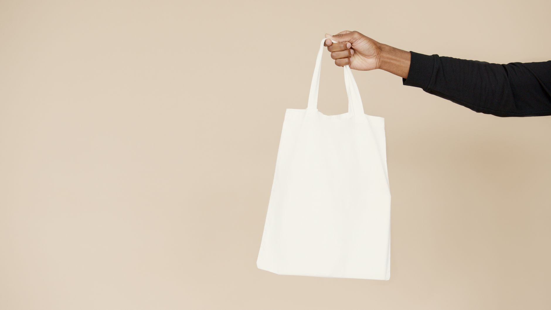 En hand som håller i en tom, vit tygpåse mot en beige bakgrund.